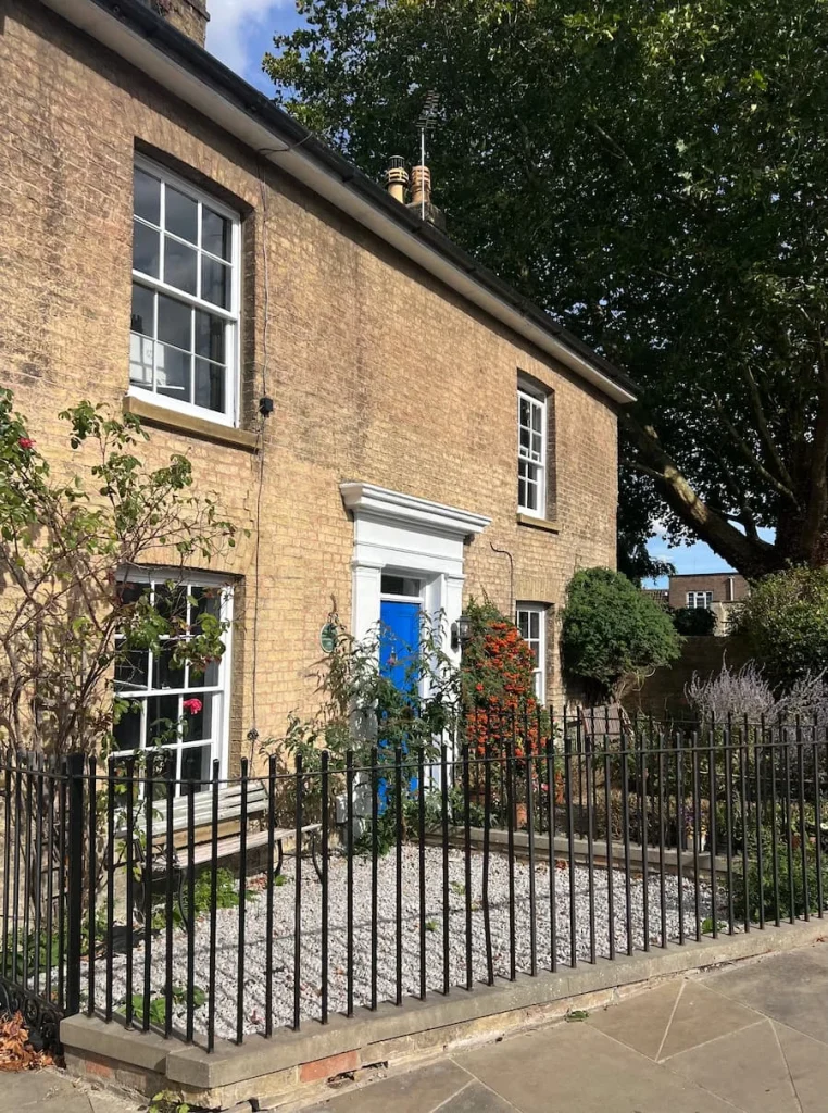 Pretty house with a blue door, Ely. There is a small garden at the front with pink flowers and a black iron gate surrounds the exterior