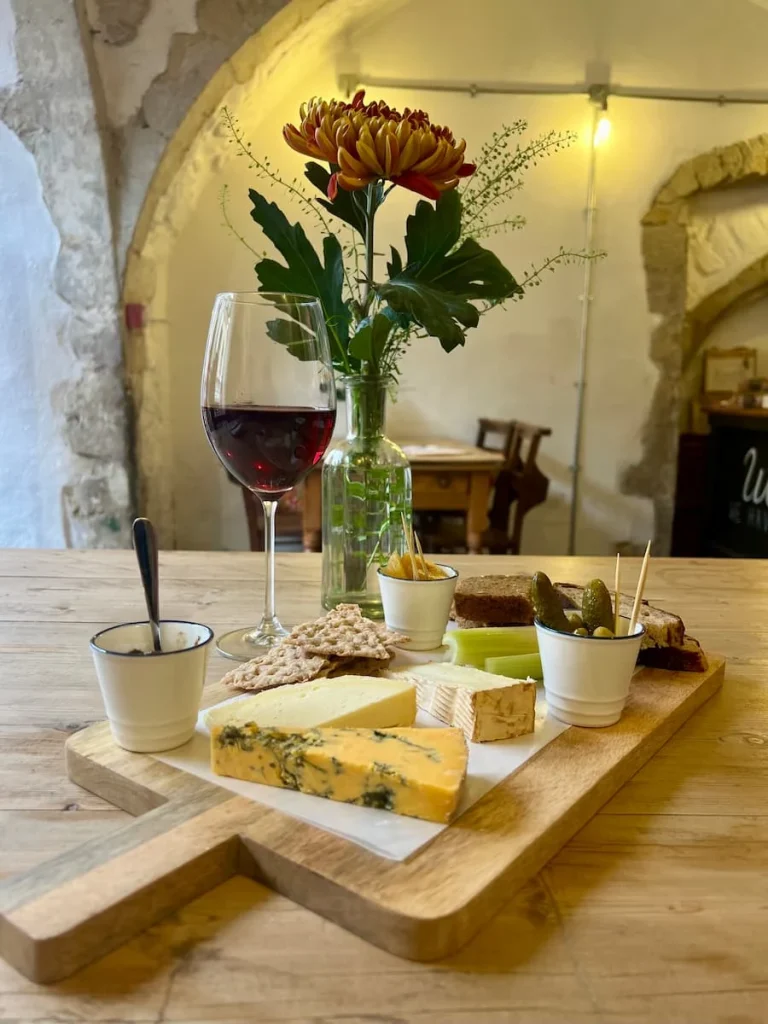 Red wine and cheese platter side view at Victoria's Cheese, Ely. There are three cheeses on the board with celery, crackers, rye bread, gherkins, chutney and red wine at the side