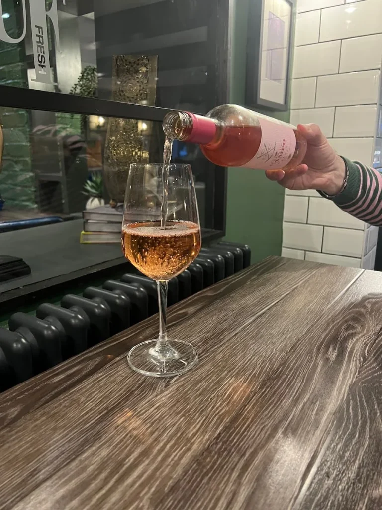 Rose wine being poured at Urban Fresh, Ely. The wine glass is on a dark brown wood table. The hand pouting it has a green and pink striped sleeve