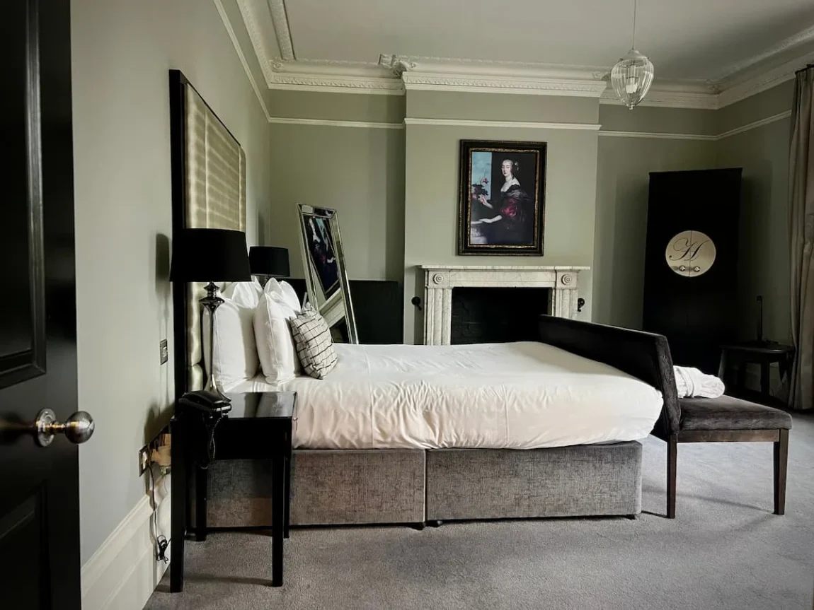 Side view of Suite 5 at Poet's House Hotel, Ely. The image shows the bed made up from a side view plus teh fireplace, artwork, dark wood wardrobe and freestanding silver framed mirror. There are also 2 bedside tables with black shaded lamps.