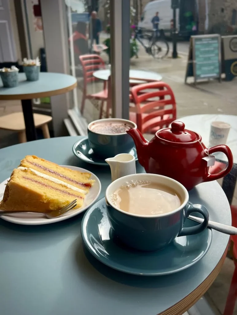 Side view image of a red teapot, Victoria Sandwich slice, tea in a grey cup and a hot chocolate in a gre cup. The items are on a blue table top with a small container of milk at Tom's Cafe in Ely