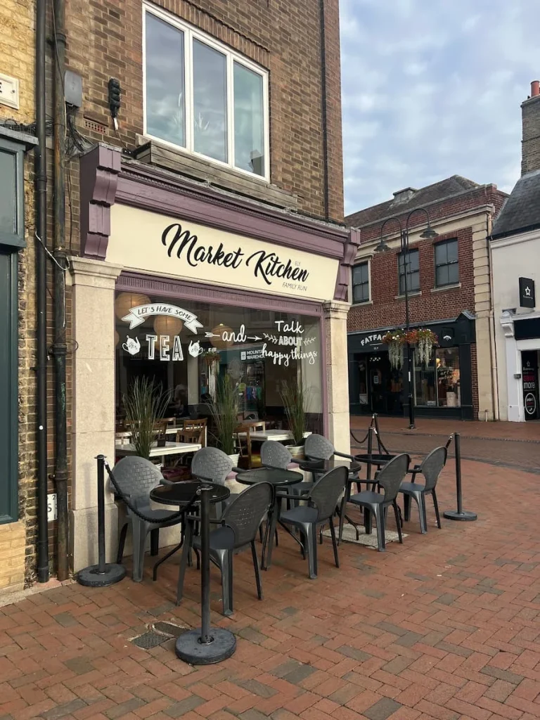 Side view of the exterior of Market Kitchen, Ely. The exterior is cream with black writing and there are chairs putside with bistro tables