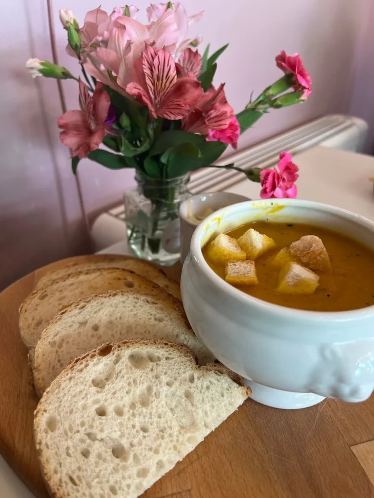 orange Soup with Croutons in a white soup bowl and slices of bread at Ely Grazing Company. There is a small vase of pink flowers on the small chopping board.