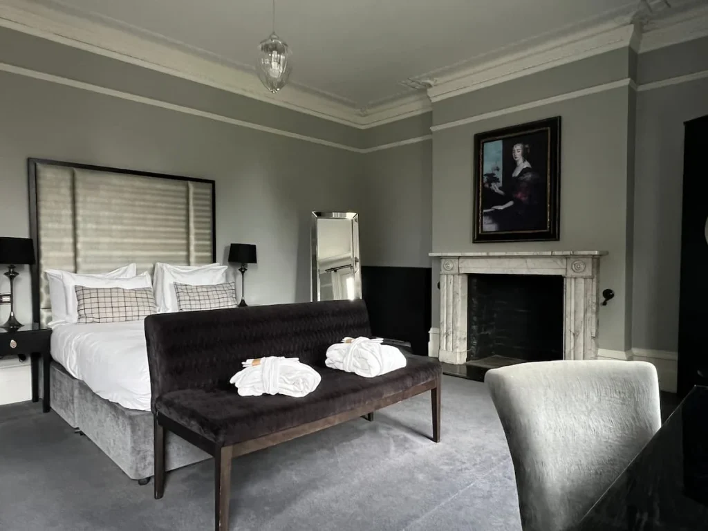 Suite 5 at Poet's House Hotel in Ely. A velvet black sofa seat with dressing gowns and a tall white headboard with crisp white bedding and grey scatter cushions. There is a ornate fireplace on the left above which is an old painting of a lady. There is also a freestanding silver framed mirror