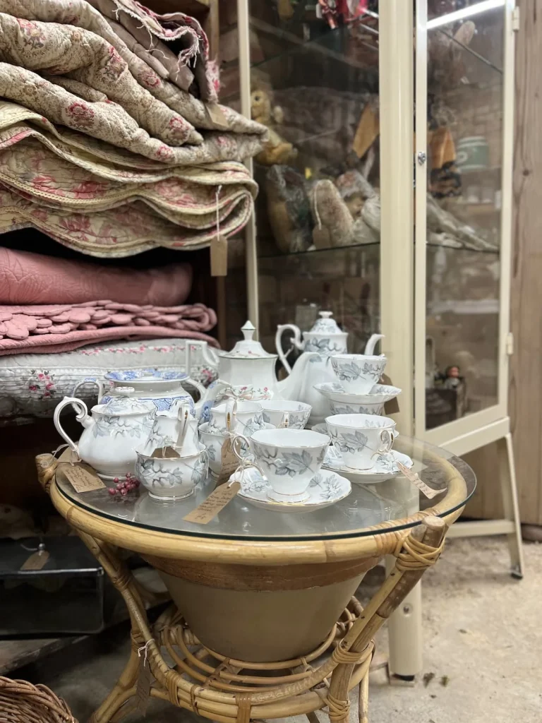 A small circular table with vintage china teapots and tea sets with table cloths in the background at Waterside Antiques, Ely