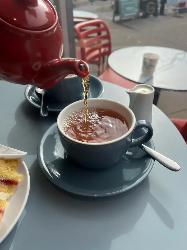 Tea being poured from a red teapot into a grey cup with saucer on a light blue table at Tom's Cafe in Ely