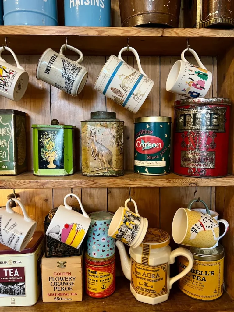 Teacups hanging on a side board with jams and conserves at Peacocks Tearooms, Ely. There are mugs and tea cups hanging on hooks on a brown wooden sideboard which also has teas and conserves on its shelves