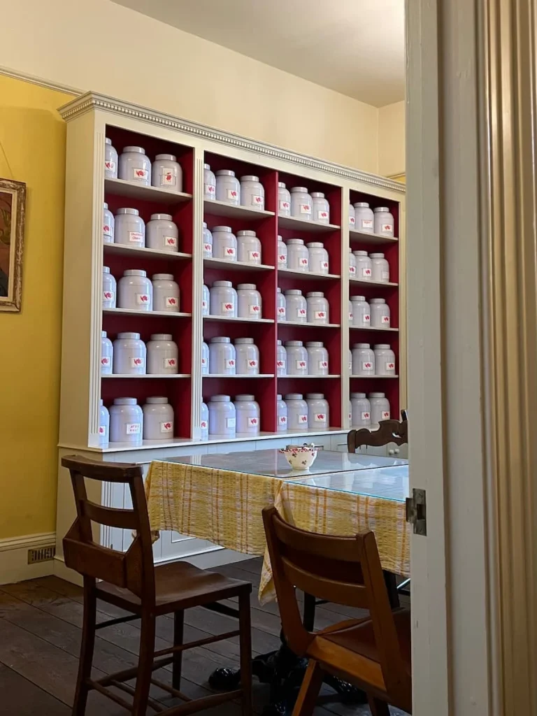 Teas in white jars at Peacocks Tearooms, Ely. The jars are arranged in order on a shelving unit with 3 white jars per section. There is also a table with brown dark wood chairs in front