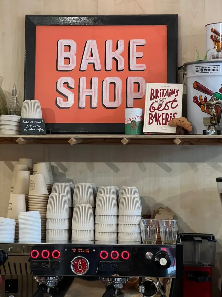 The Bake Shop sign with pink letters and a red background at Bake Shop General Store in Ely. The shelves surrounding hold cups and other cafe wares.