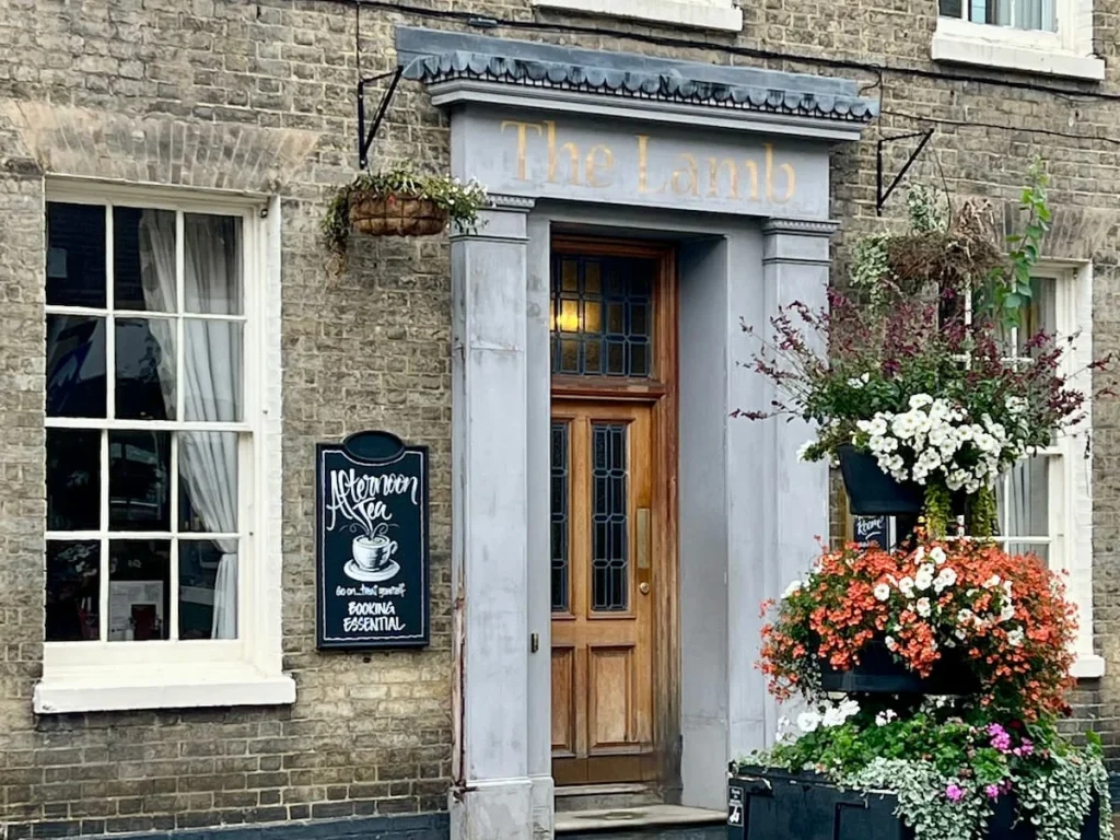 The entrance of The Lamb Hotel, Ely exterior with a light brown wooden door and floral arrangements. There is a chalk board advertising an afternoon tea on the outside of the building.