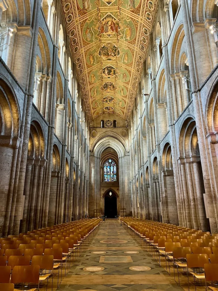 A view of the Nave in Ely Cathedral looking down towards the front entrance. There are chairs on either side with a painted ceiling above and white arches down either side.