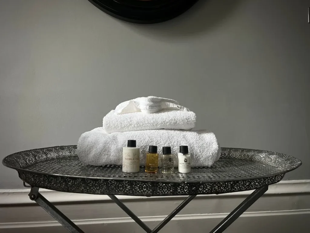 A pile of different towels at Poet's House Luxury Boutique Hotel in Ely. The towels are placed on a silver oblong metal table and in front of them are 4 bottles of moisterizer, shampoo, conditioner and bath gel. the wall behind is light grey.