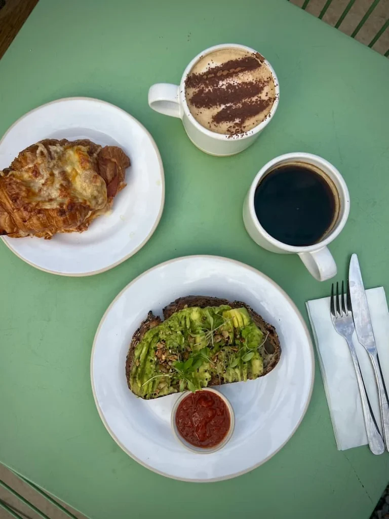 Top lay of brunch at Gail's Bakery, Ely. Avocado topped sourdough with chilli sauce, cheese croissant, cappuccino and a black coffee on a pale green table top.
