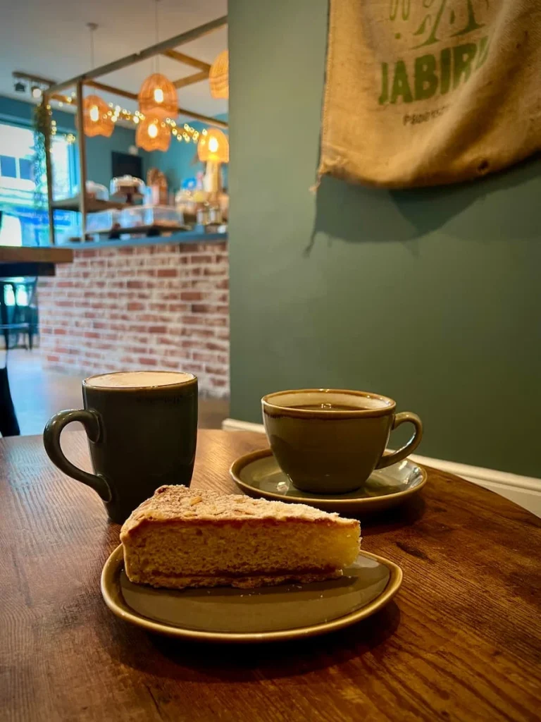 Side view of a chai latte, black coffee and slice of Victoria Sandwich Cake on a brown table with the coffee counter and a teal wall in the background at Tamarin Bay Coffee House, Ely