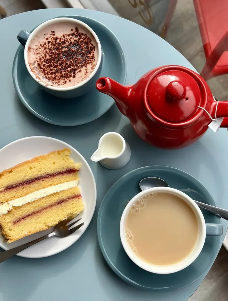 Top lay image of a red teapot, Victoria Sandwich slice, tea in a grey cup and a hot chocolate in a gre cup. The items are on a blue table top with a small container of milk at Tom's Cafe in Ely