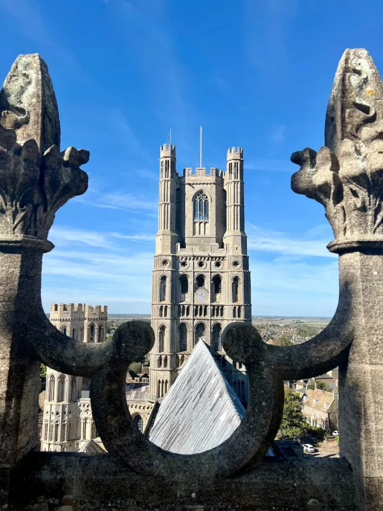 Tower from the roof of Ely cathedral against a bright blue sly with an inter connecting slate roof top between teh two towers. In the distance there are Ely landmarks