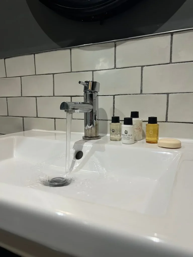 Bathroom white sink at Poet's House Luxury Boutique Hotel in Ely with tap running and small bath product bottles to the side. The back wall has white brick-style tiles and the wall paint is dark grey