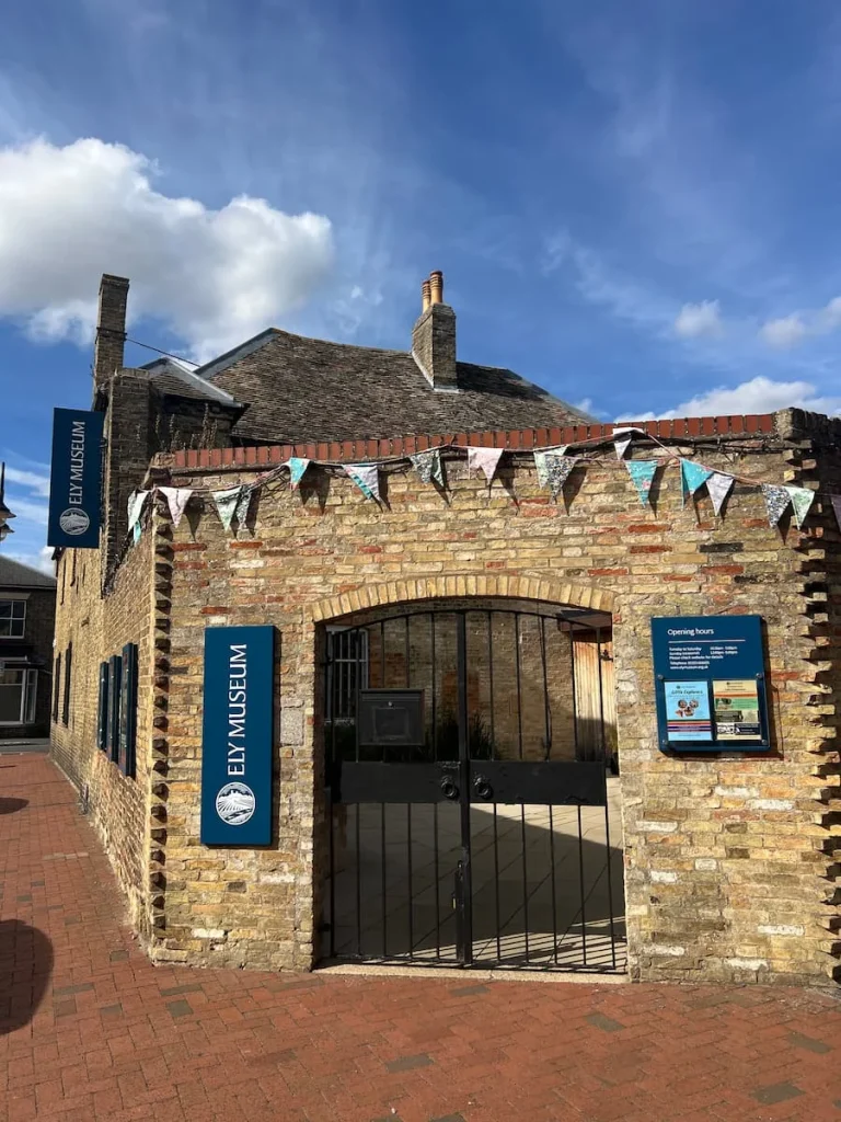 The brick exterior of Ely Museum with teal coloured signs at either side of the main wrought iron gate entrance. There is pale pink and blue bunting on the wall above teh gate.