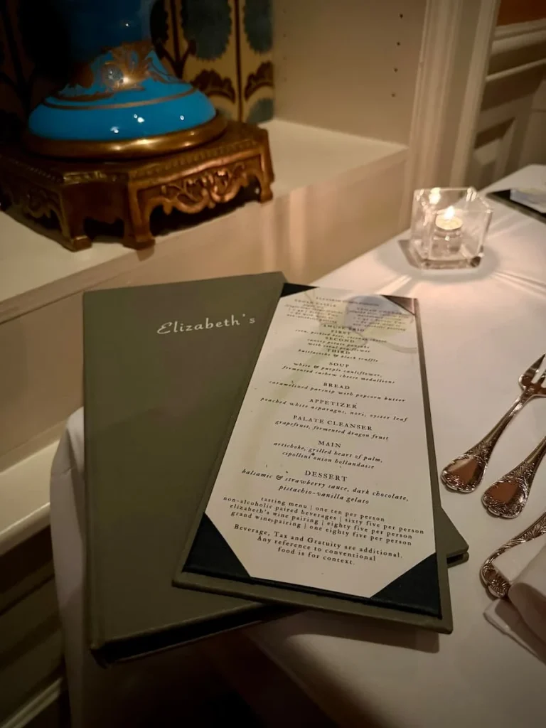 10 course menu on table with cover at Elizabeth's Washington DC. There is a blue and gold ornamental vase on teh table and a small candle light under the white table cloth