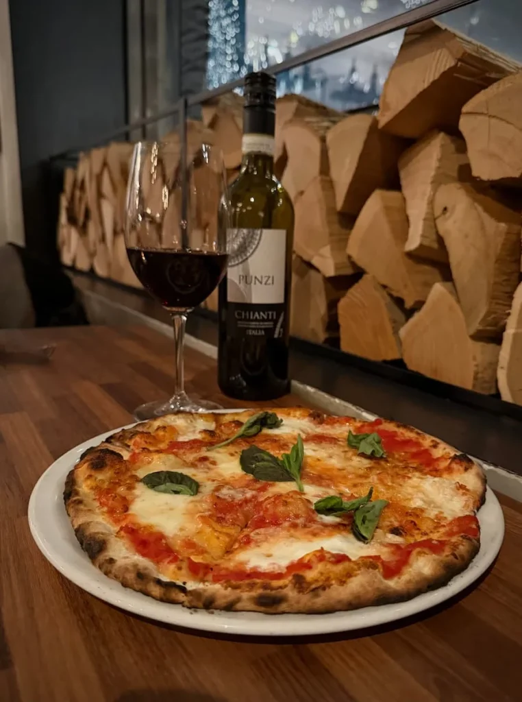 A small margherita pizza with a well done crust witha glass of red wine and Punzi bottle in the background at Via Sophia, Washington DC. There are wooden cut logs in the background