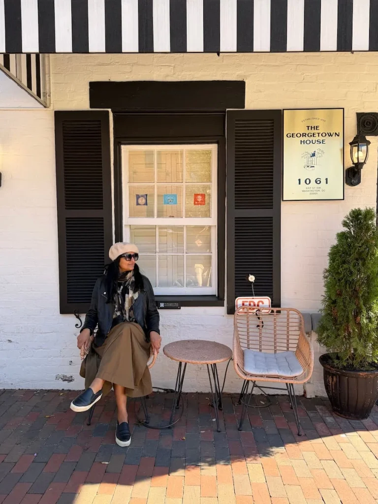 Bejal sitting outside shop on C&O Canal, Georgetown, Washington DC on a chair with a black leather jacket, light brown skirt, cream beret. There's a window in the background with brown shutters.