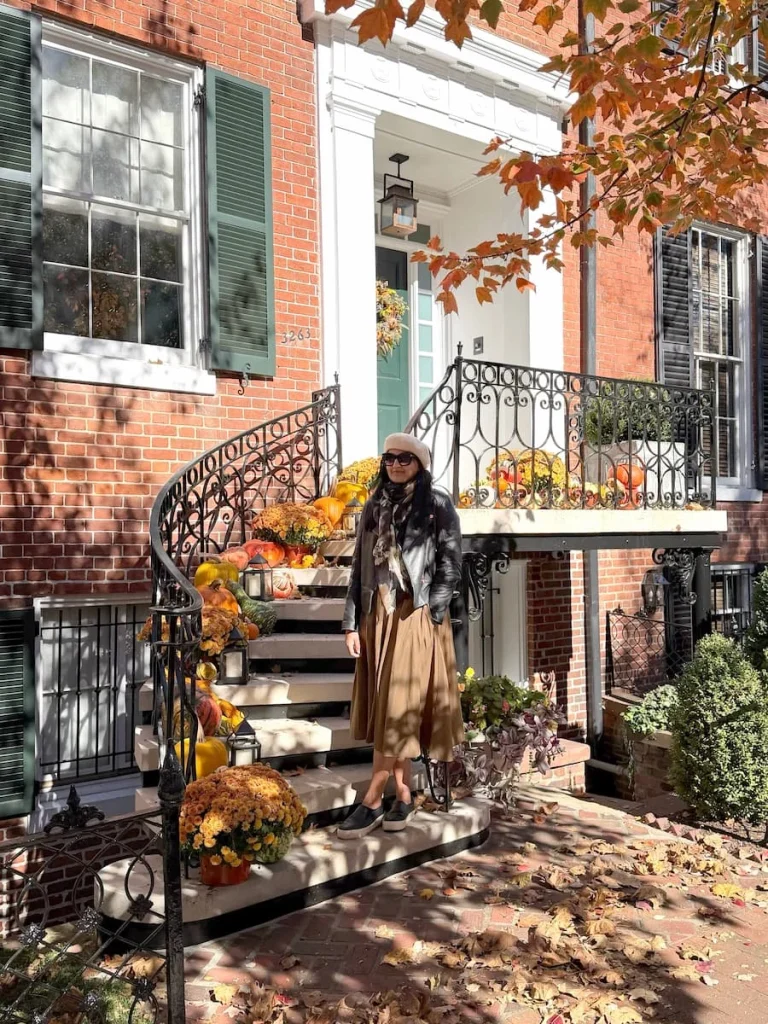 Bejal standing at the bottom of a curved wrought iron staircase in Georgetown Washington DC. There are pumpkins on the staircase. Bejal is wearing a cream beret with black leather jumper and a brown skirt