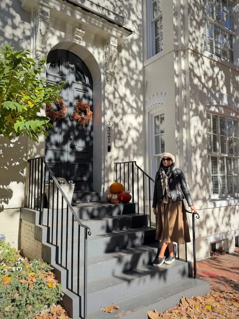 Bejal standing outside houses decorated for Autumn in Georgetown Washington DC. Bejal is wearing a black leather jacket, brown skirt and cream beret and black pumps.