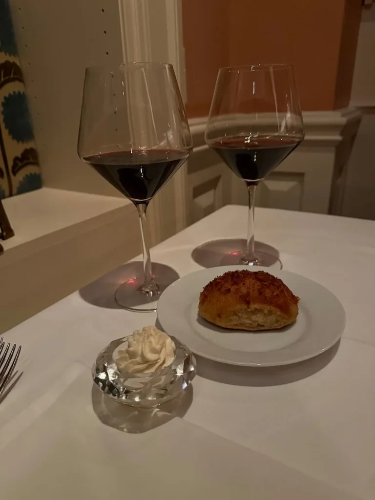 Bread course at Elizabeths, Washington DC. Caramelized parsnip with popcorn butter in a small glass bowl and a bread roll on a white plate. There are 2 red wine glasses in the background.