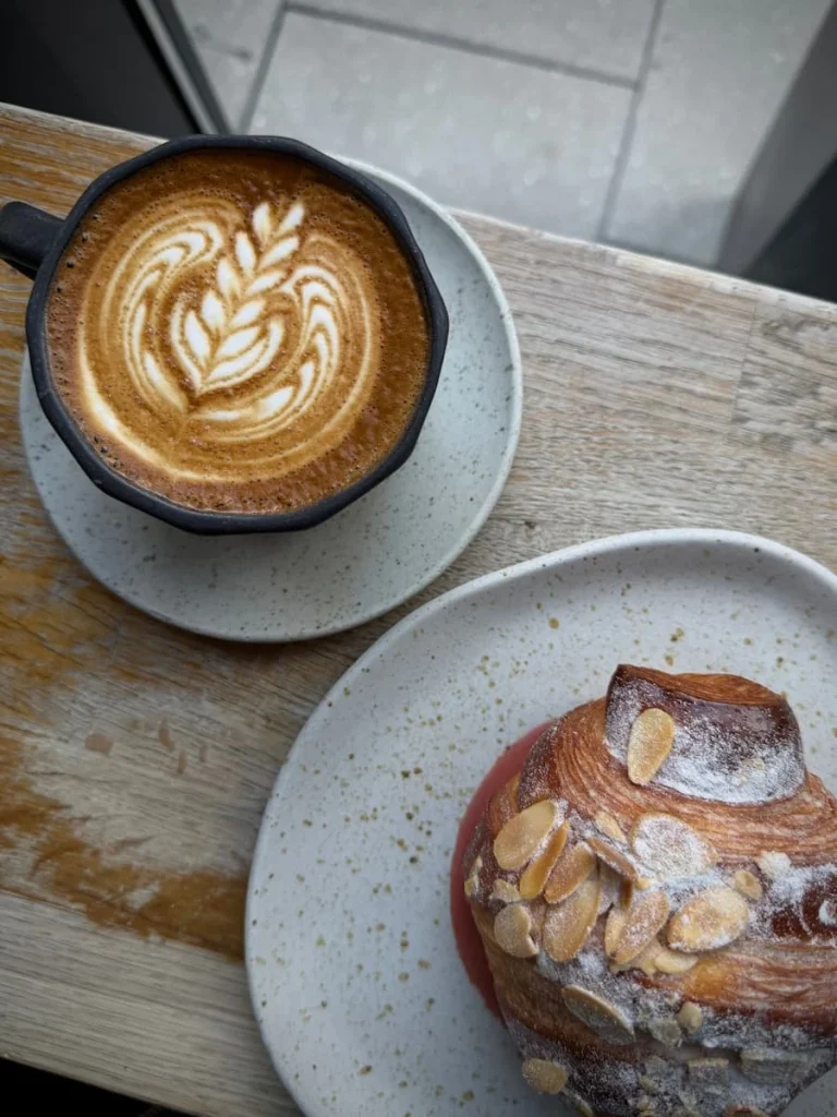 Flat lay of a Cappuccino and almond pastry at Dua DC Coffee, DC. The table top is of a light wood colour and the cappuccino is in a dark grey earthenware cup and the almond croissant is on a white earthenware side plate. The cafe is located in Washington DC City Centre.