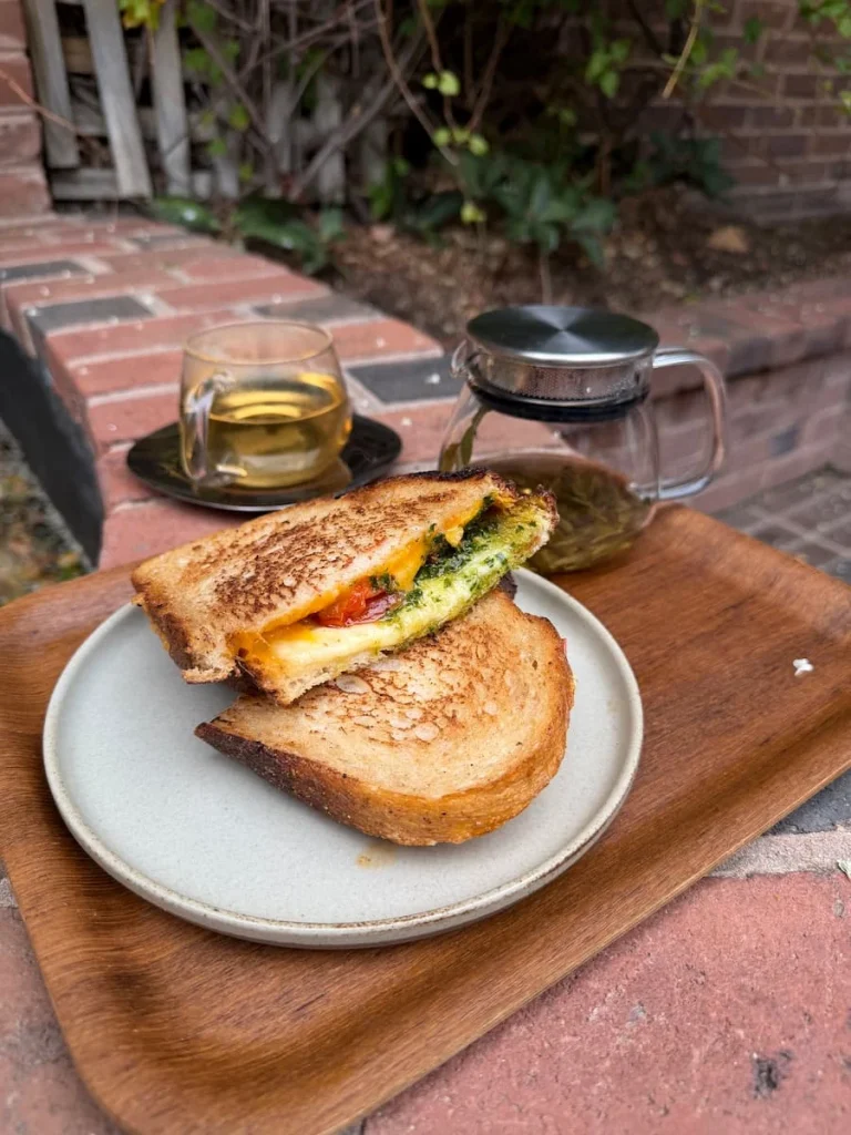 Cheese, tomato and pesto toastie on toasted sourdough on a cream plate with a coup of jasmine tea and jasmine pearls in a glass pot. The brown tray is on a brick wall at the Flor courtyard in Georgetown, Washington DC