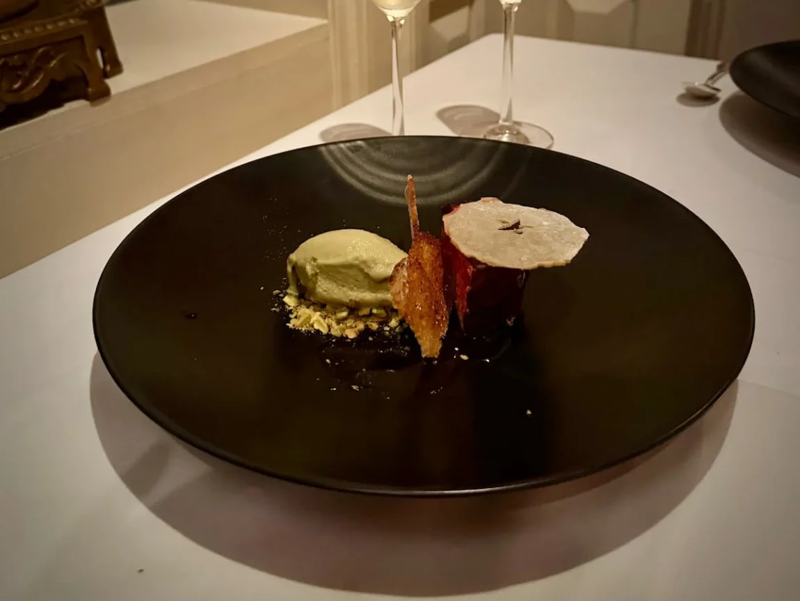 Dessert at Elizabeth's vegan fine dining in Washington DC. A large black plate with balsamic & strawberry sauce, dark chocolate, pistachio-vanilla gelato. There are two flutes of soarkling wine in the background against the salmon pink wall and blue antique vase. Image is in portrait format.