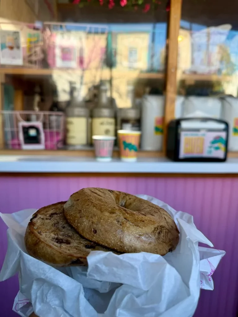 How to spend One day in Georgetown Washington DC. Bejal holding a cinnamon and Raison bagels at Call Your Mother, Washington DC. The bakery counter is in the background with the coffee and napkin dispenser and pink counter