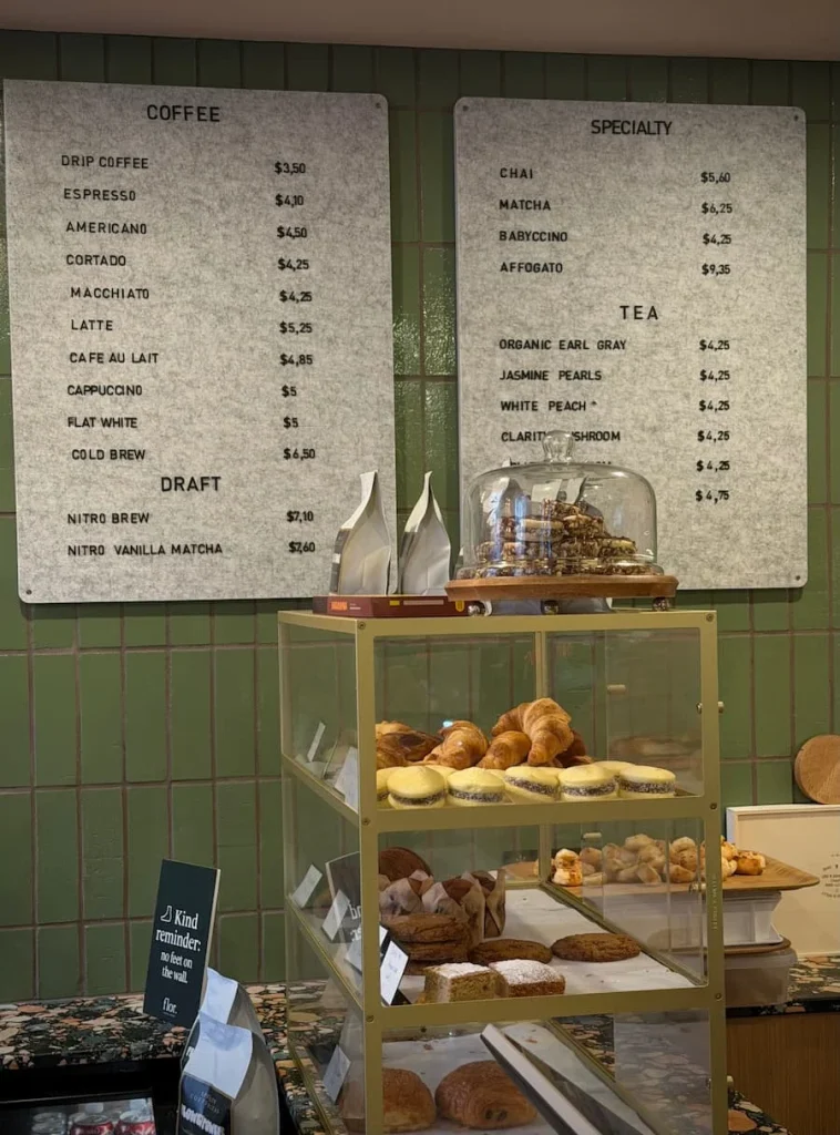 Coffee counter at Flor, Washington DC. There is a small glass cabinet with pastries and cakes behind which there is a board with drink opetion on the light sage coloured wall of the cafe in Washington DC.