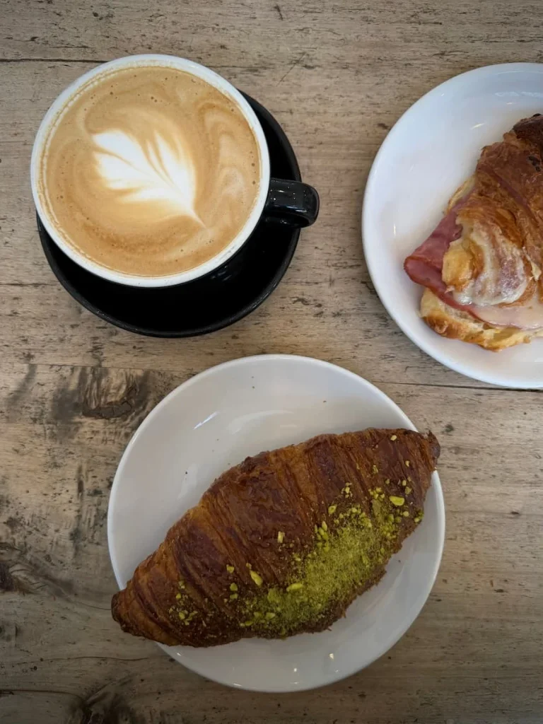 A flat lay of on a light oak table top of a pistachio croissant, cheese croissant and cappuccino in a black mug at Tatte in Washington DC