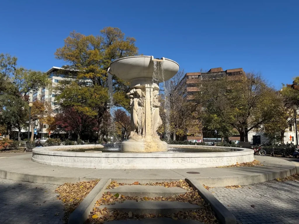 Dupont Circle Fountain with foliage all around and people sitting on benches at midday. Dupont Circle has a few sustainable hotels in Washington DC