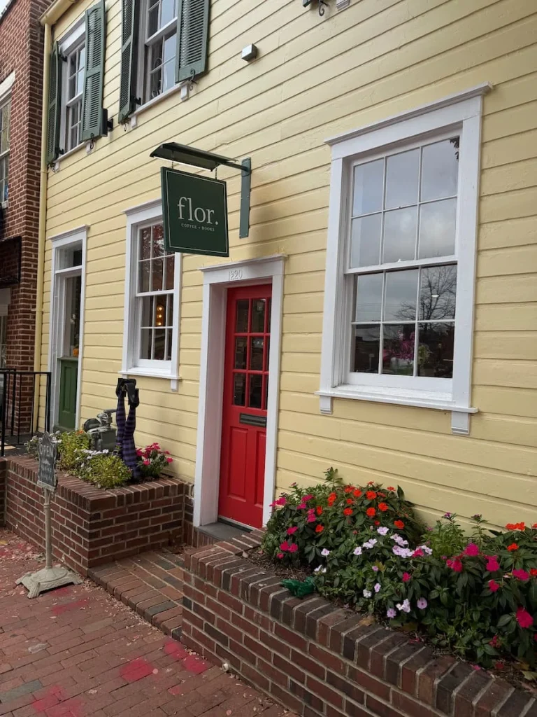 Exterior of Flor, Washington DC. The exterior has a crea, wooden finish and a red door with a hanging sign reading, Flor. There are some small pretty flowers on the floor around the cafe in Georgetown, Washington DC.