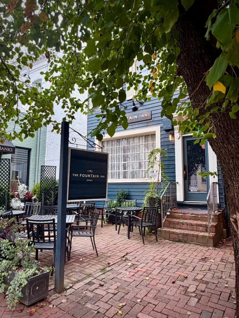 Exterior of Fountain Inn in Georgetown Washington DC. The grey exterior has a black sign hanging outside it with a large tree with green leaves covering the courtyard area