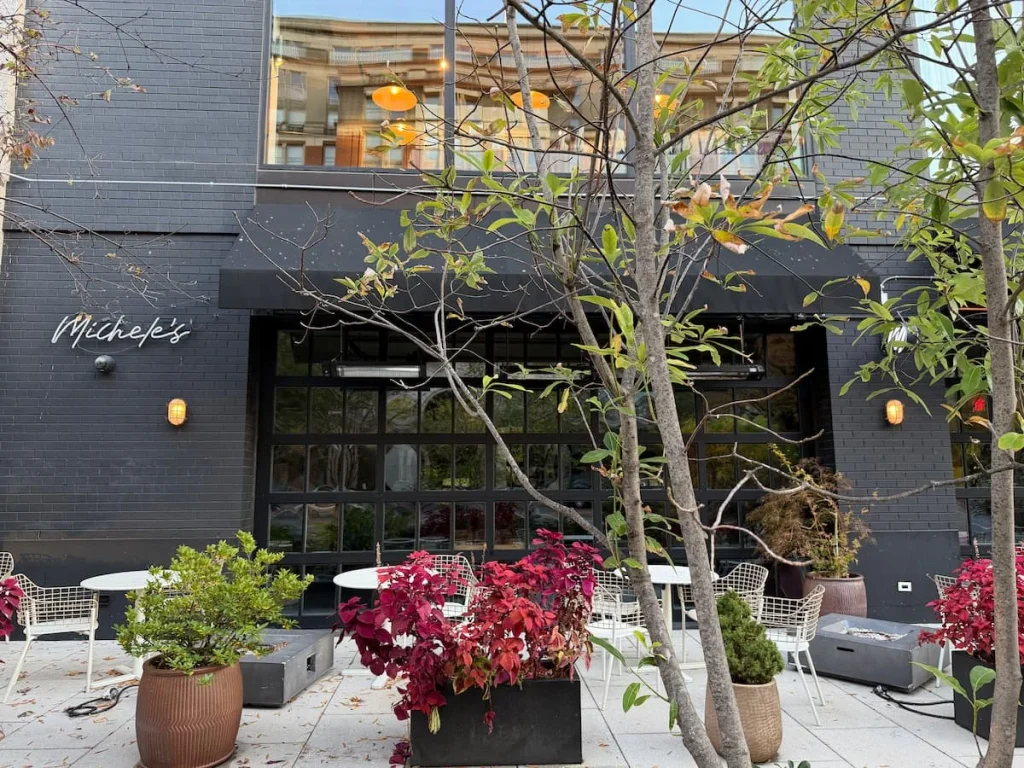 The exterior of Michele's in Washington DC with red and natural leaved plants in pots and foliage around teh exterior windows