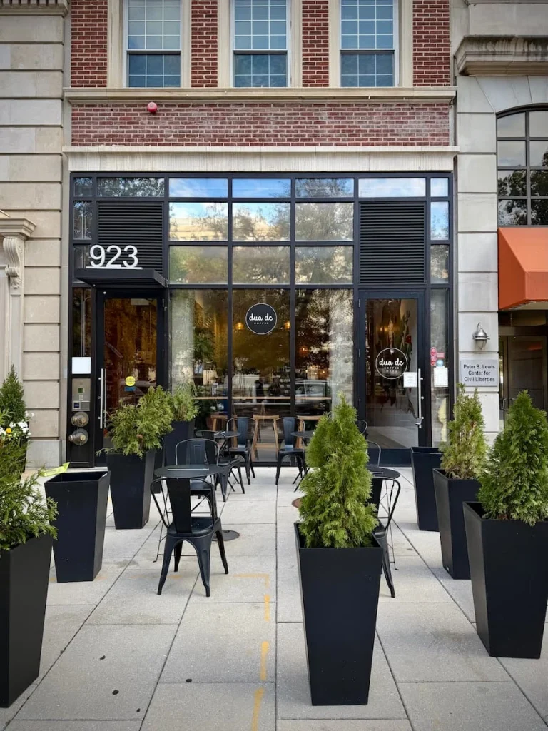 Exterior of at Dua DC Coffee, in Washington DC. There are tall black pots containing foliage with the coffee shop from and bistro tables and chairs in the forecourt of the shop. One of the cafes to enjoy Vegan and Vegetarian food in Washington DC