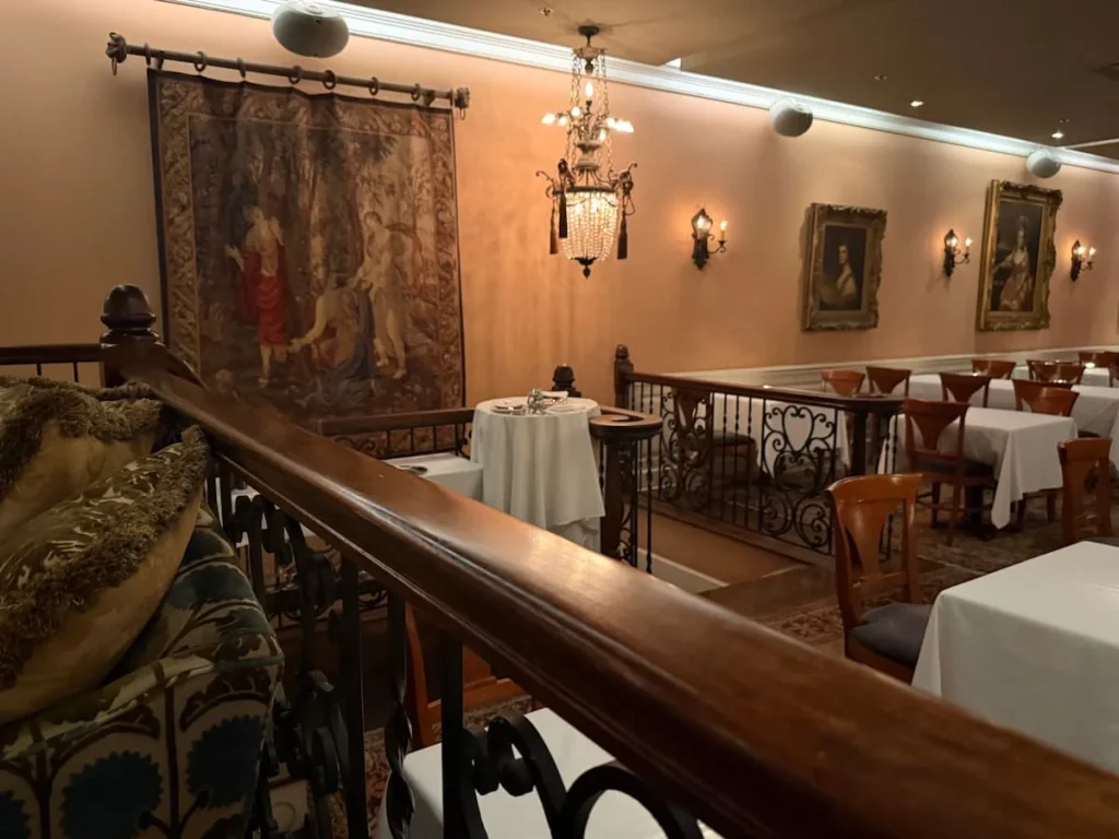 First floor seating for diners at Elizabeths, Washington DC. There are 6 tables with white table cloths and cutlery. The walls are a pale pink colour with photos on the wall with brown and antique gold frames. The lighting is low. The image format is portrait