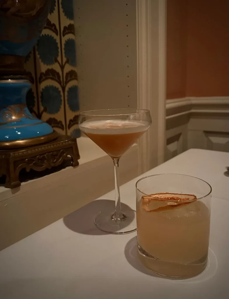 A Hemingway in Havana and Amaretto ginger Sour at Elizabeths, Washington DC under a white table cloth. There is a blue ornate vase to left with a salmon pink wall to the right of the image