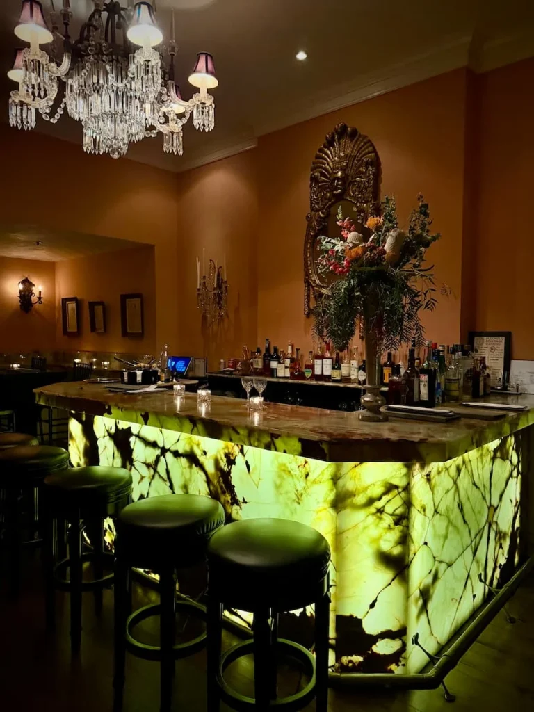 Onyx bar at Elizabeth's, vegan fine dining in  Washington DC. The exteroor of teh bar is lit up with speckled green adn black under lighting and the wall behind it has a ornate baroque framed mirror with a flower vase. The bar area has a row of neatly arranged spirots and there are a few place setting in the bar counter with high top stools.