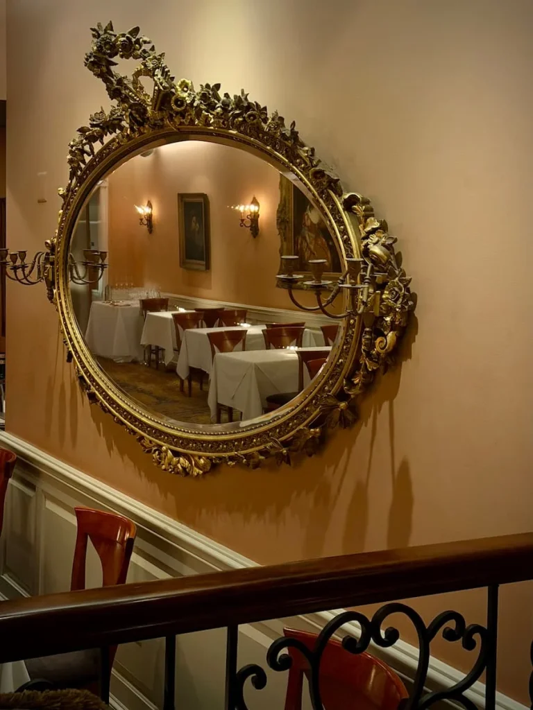 Ornate Baroque framed mirror with reflection of seating on the first floor. The wall is painted a cream/pale pink and there is a dado rail underneath