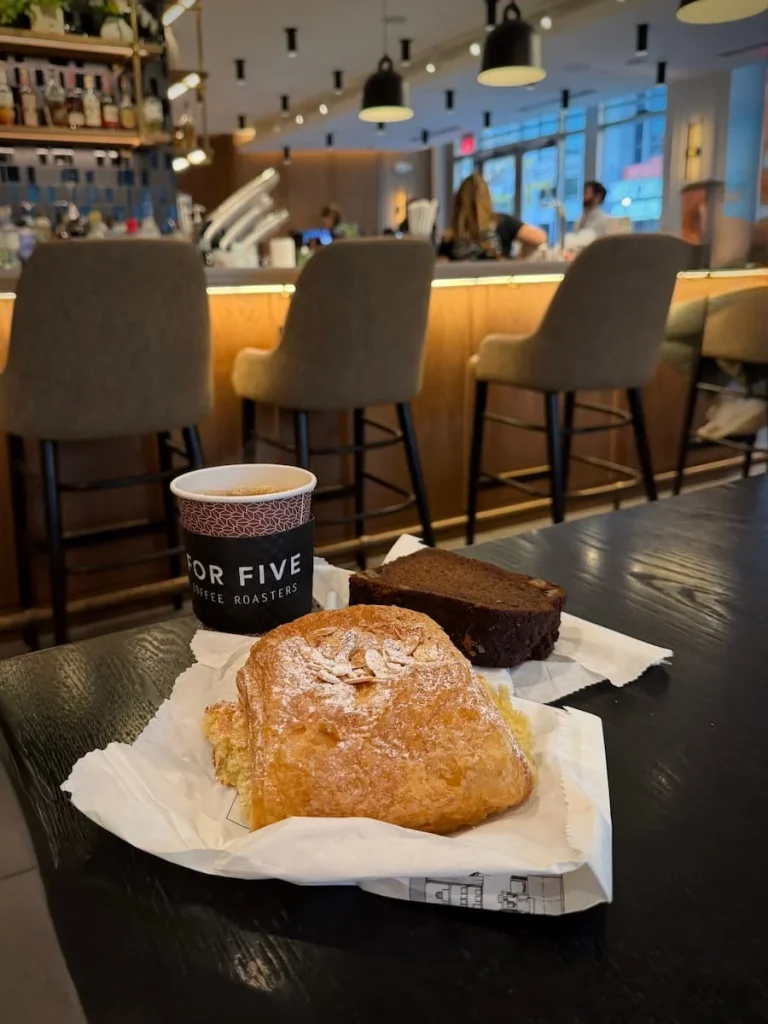 Pastries and coffee at For Five Coffee, Washington DC. On a black table top there is an almond croissant and a banana and walnut slice with a cappuccino. The main bar is in the background.