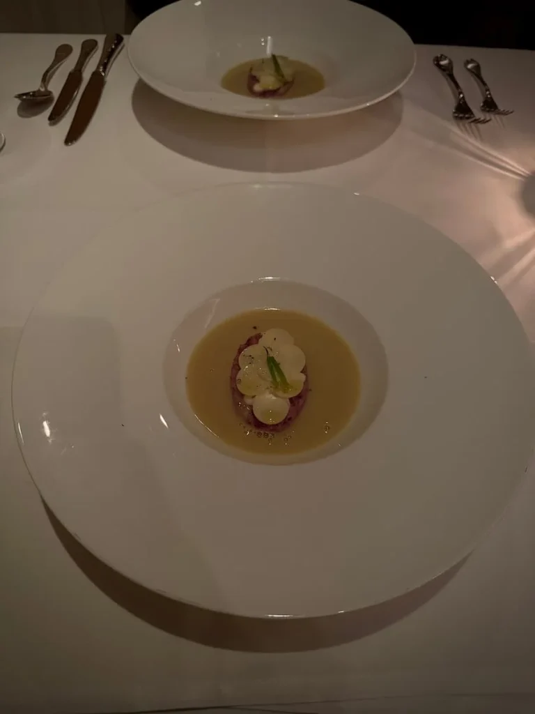 Soup dish at Elizabeth's, Washington DC. The bright white large plate has a yellow coloured soup in the small circular indent in the middle. The same dish is mirrored with cutlery at the top of teh image for the otehr table setting
