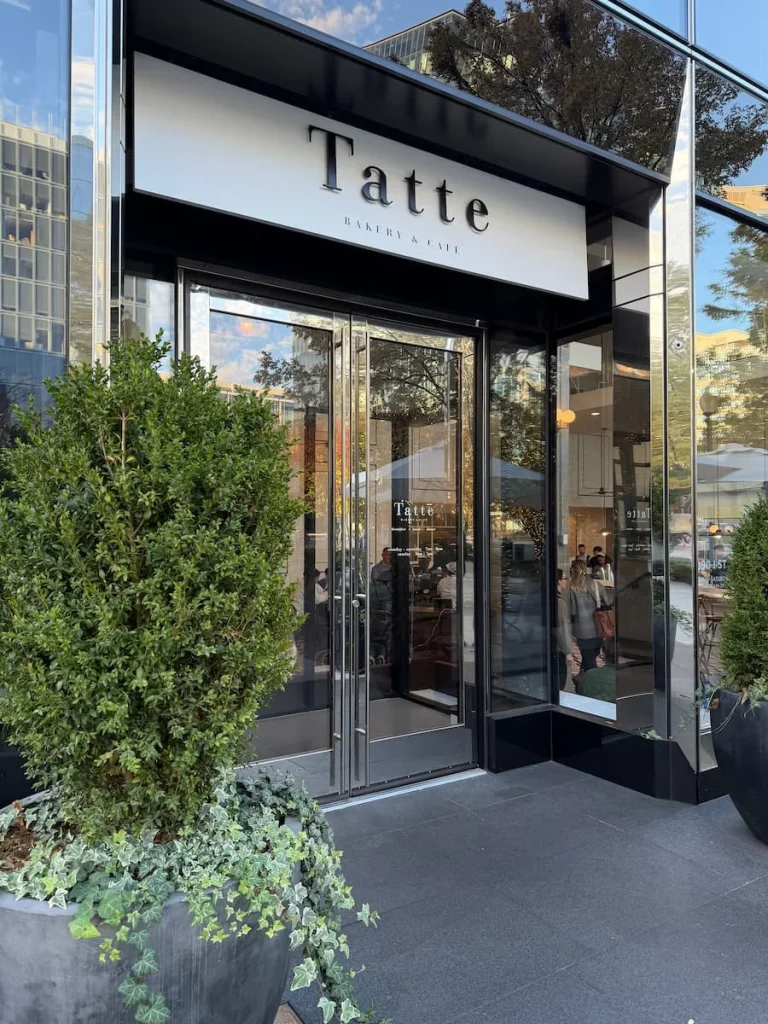 The glass exterior of Tatte with plants in the entrance and a black sign against a white background in Washington DC