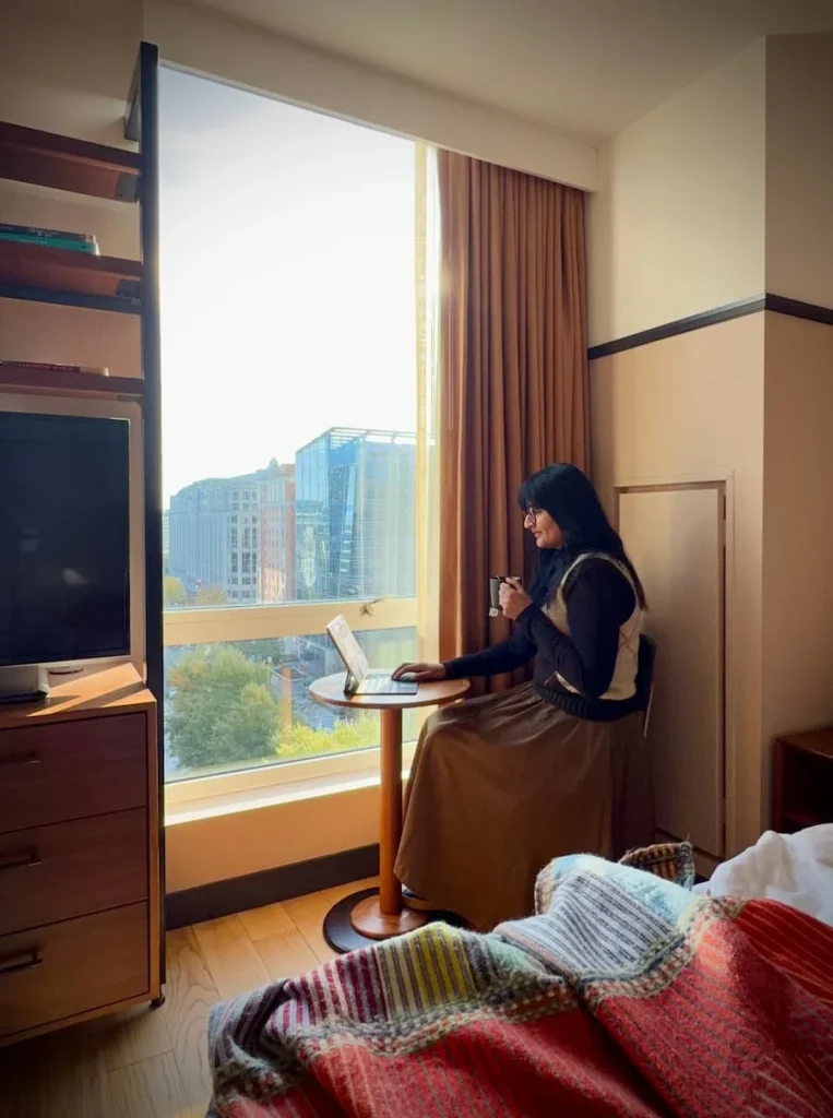 Bejal sitting on bed working on laptop at Eaton DC, Washington DC. There are street views from the windoe behind Bejal