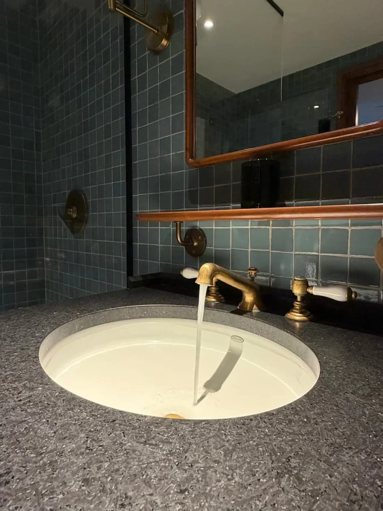 Tap with running water in vanity basin at Eaton DC, Washington DC. The tap is gold and there is blue/teal tiling in the background against dark wood work