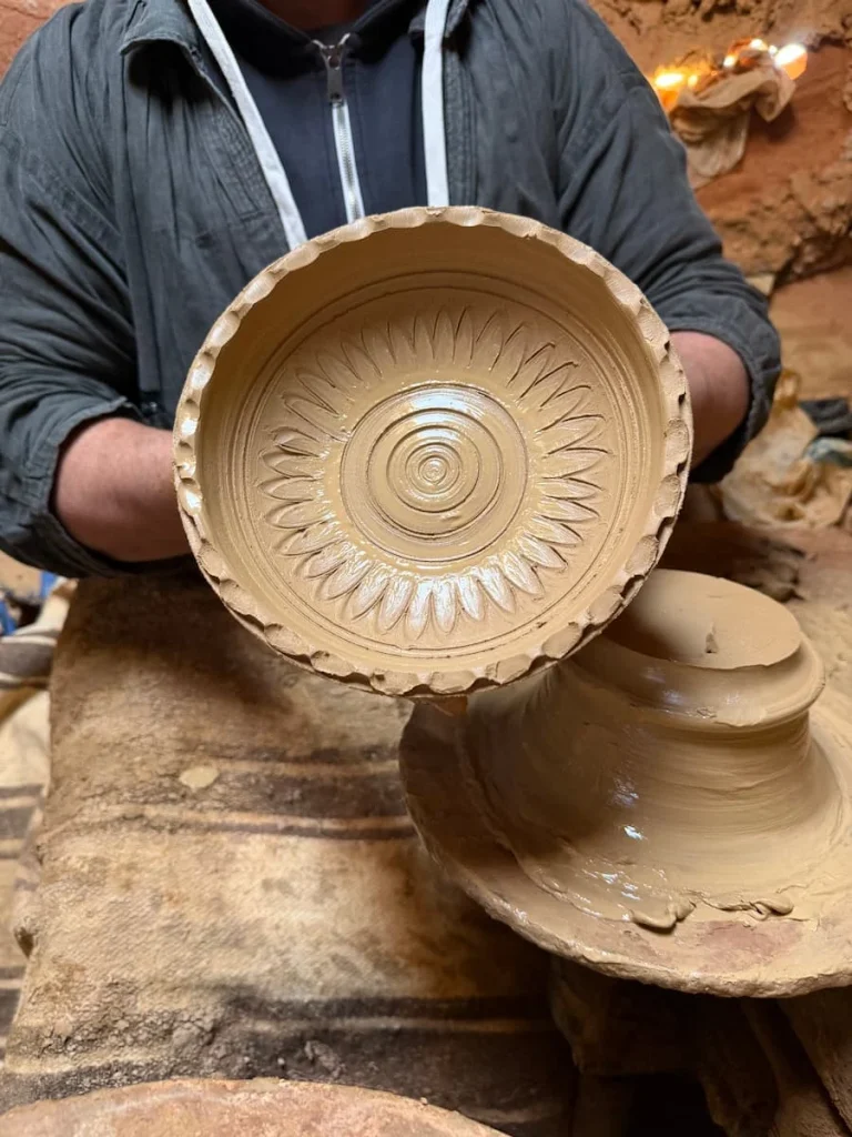 A clay ash tray with designs which has just come off the wheel. The potter is holding it up with his wet hands and is wearing a grey hoodie. The ash tray design is circular with lines coming out from the centre to the outside