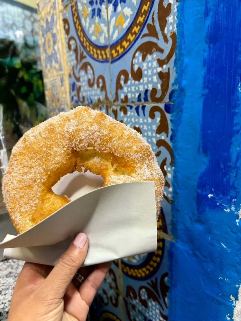 Bejal holding a Bamboulini in Sidi Bou Said, Tunisia, against a blue mosaic door. The Bamboulini is a deep fried doughnut rolled in sugar.